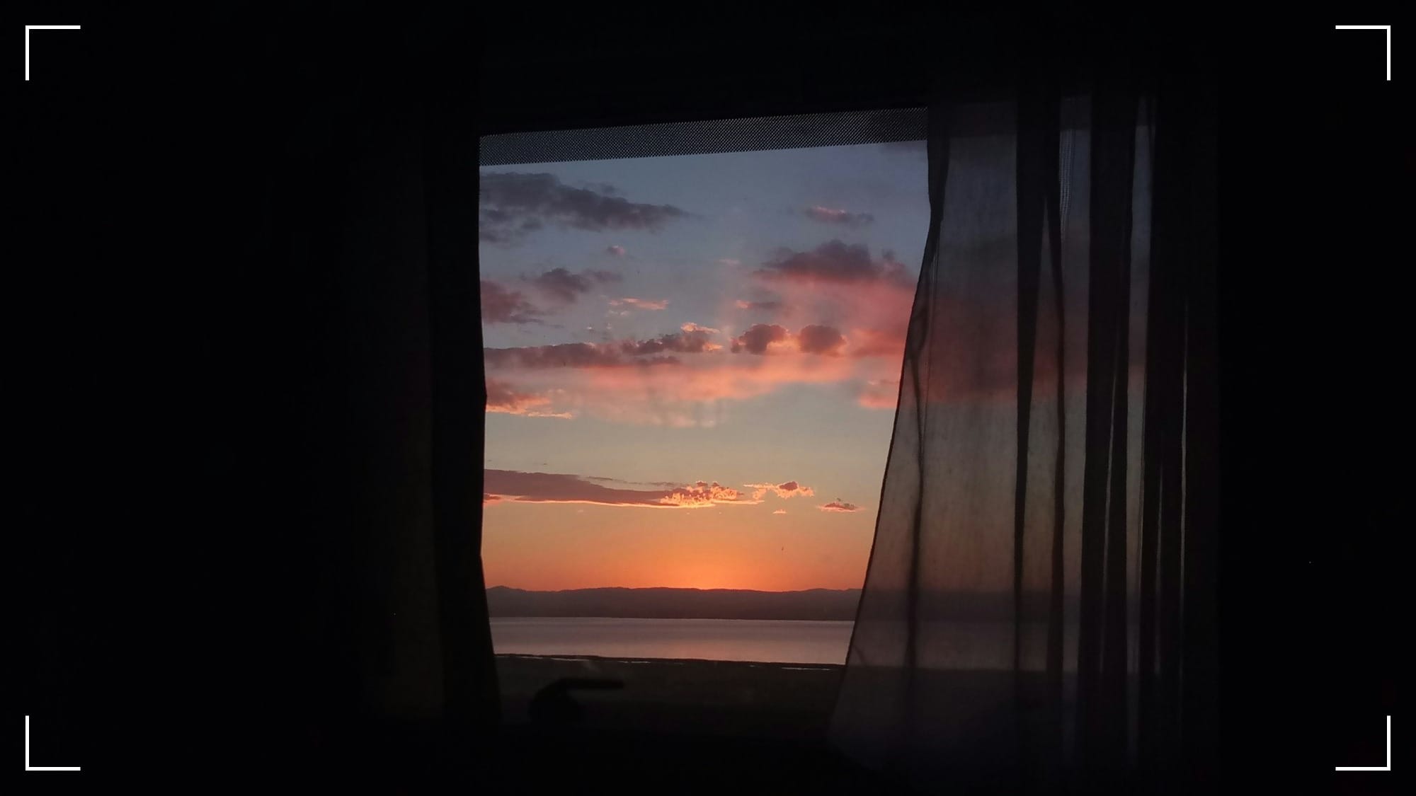 A view from a dark room looking through a window at a vibrant sunrise over water. The light of dawn breaks through the shadows, inviting the viewer to step outside into the new day.