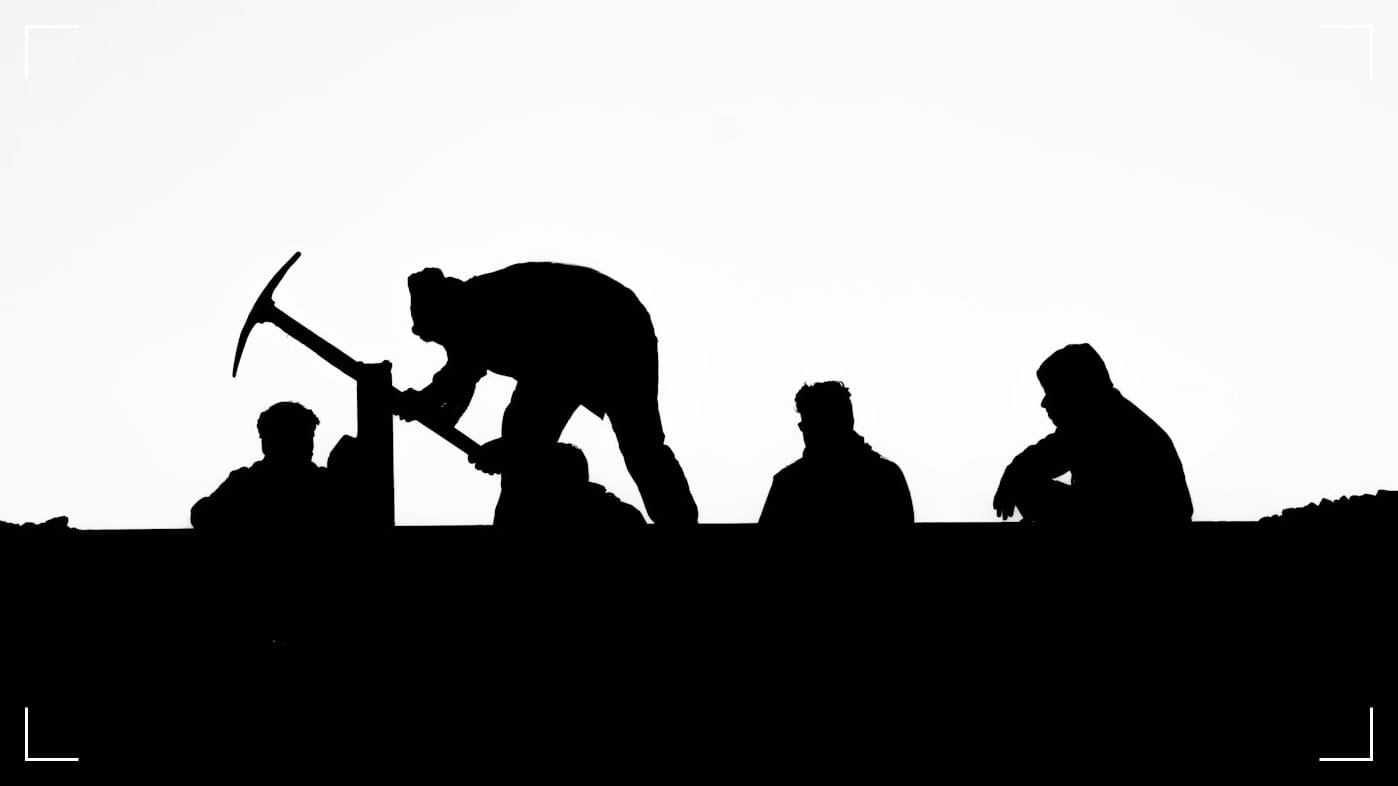 High-contrast silhouette of four laborers on a horizon line. One man swings a pickaxe while three others observe or rest nearby, set against a plain, bright white background