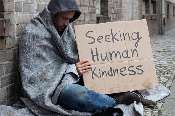 A person sits on a sidewalk wrapped in a blanket, holding a cardboard sign that reads "Seeking Human Kindness." The setting is an urban street with brick walls.