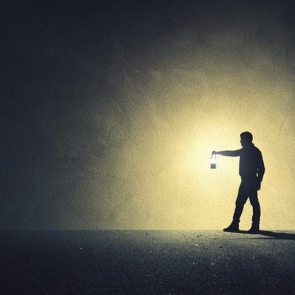 A person stands alone in the dark, holding a lantern that casts a bright circle of light around them, creating a dramatic shadow on the ground.
