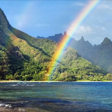 A vibrant rainbow arcs from lush green mountains down to a tropical beach, with blue ocean waves in the foreground and a partly cloudy sky above.