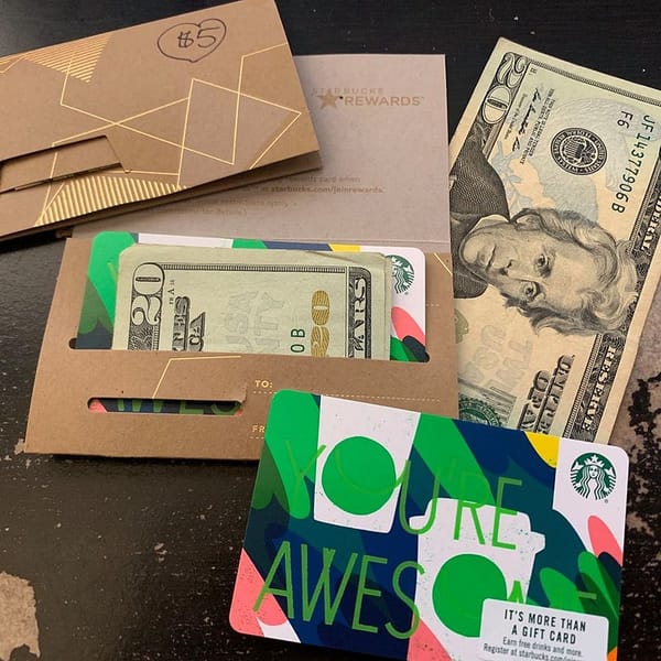 A Starbucks gift card holder with "You're Awesome" on it contains $20 bills. More $20 bills and a $5-marked envelope are nearby, along with a Starbucks Rewards card.