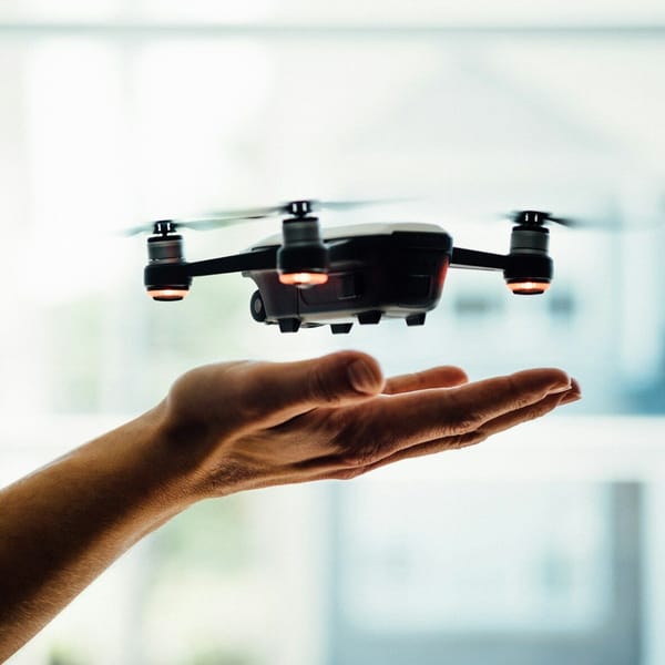 A hand is held out under a small black drone hovering indoors, with its lights on and propellers spinning.
