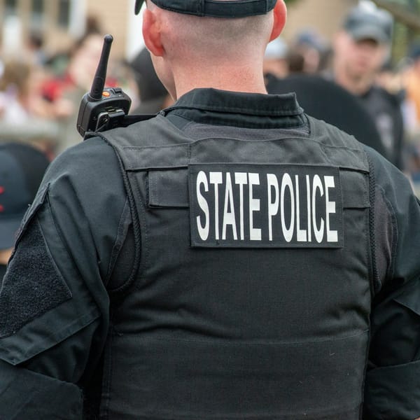 A state police officer is seen from behind, wearing a black uniform and vest labeled "STATE POLICE," with a radio on their shoulder. The background shows a blurred crowd.