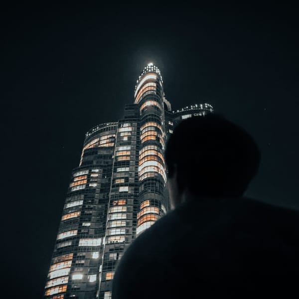 A person stands at night, gazing up at a tall, illuminated skyscraper with many glowing windows against a dark sky, creating a dramatic and futuristic city scene.