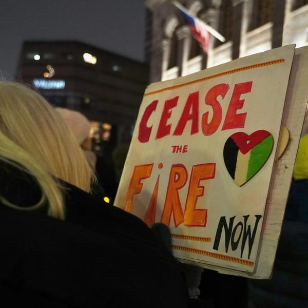 A person at a nighttime protest holds a sign reading "Cease the Fire Now" with a heart in Palestinian flag colors. Buildings and an American flag are visible in the background.